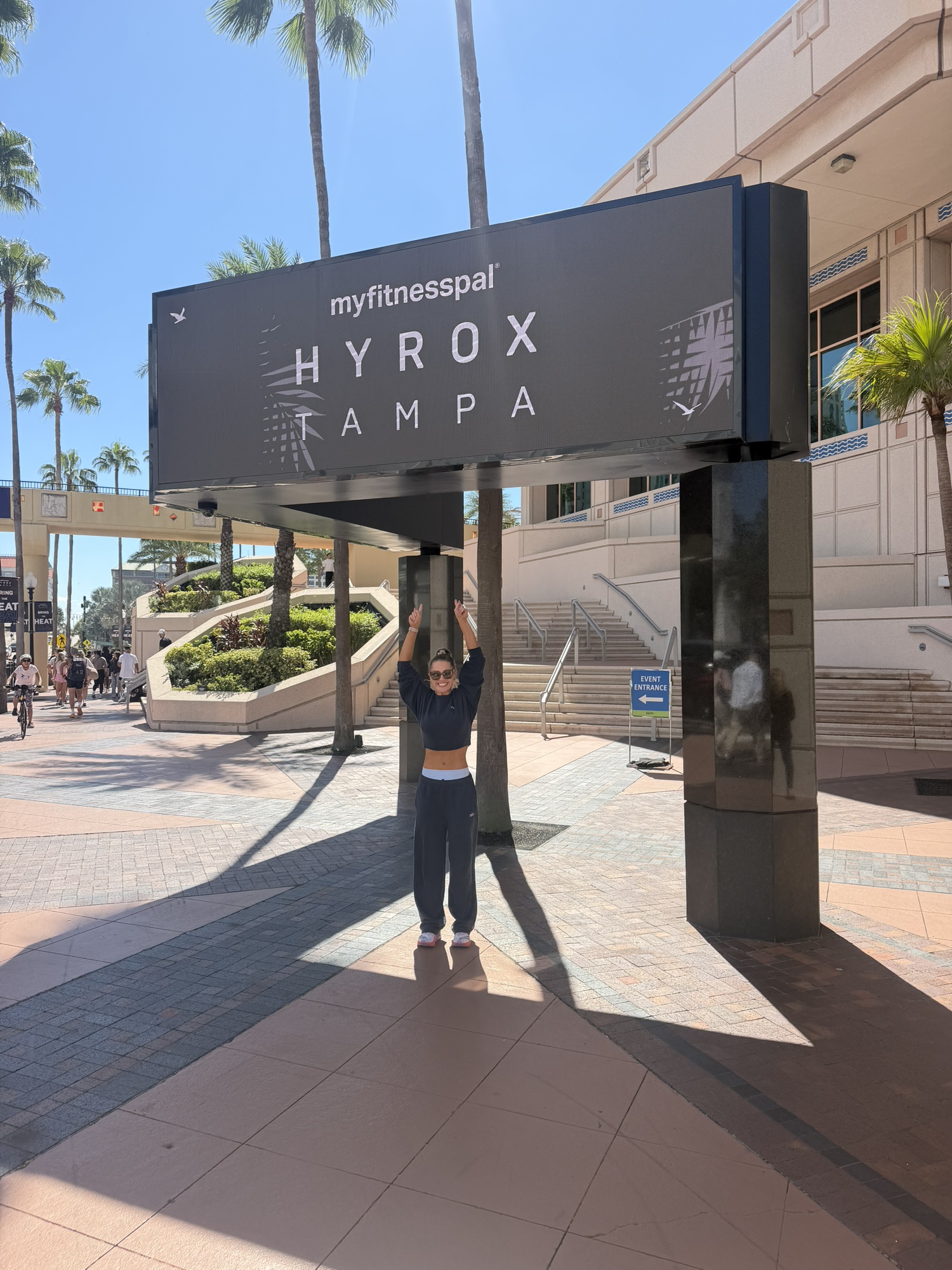 Gen under the Hyrox Tampa sign, arms up and smiling